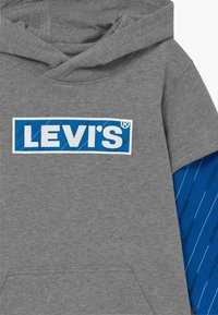 Felpa grigia realizzata in tessuto morbido con una patch blu recante il logo bianco "LEVI'S". Presenta una manica a metà con un motivo di testo ripetuto in blu.