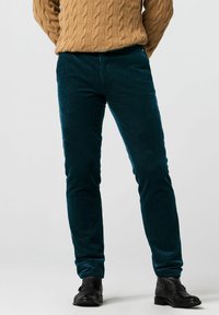 Petrolkleurige corduroy broek met verticale ribbels, slanke pasvorm, met een gladde textuur en effen voorzakken. Gedragen met een tankleurige kabelgebreide trui.