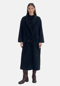 Abrigo largo negro con un diseño entallado, que presenta un cuello con muesca, dos bolsillos frontales y dos botones. Combinado con botas marrones hasta la rodilla.