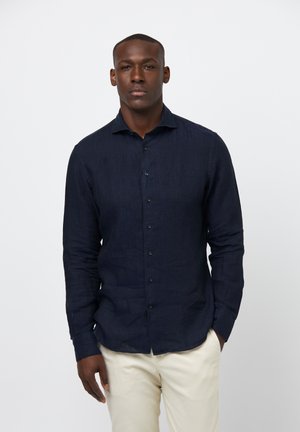 Navy blue linen shirt with long sleeves, buttoned front, and a spread collar. Worn with light-colored trousers. Smooth texture, classic design.
