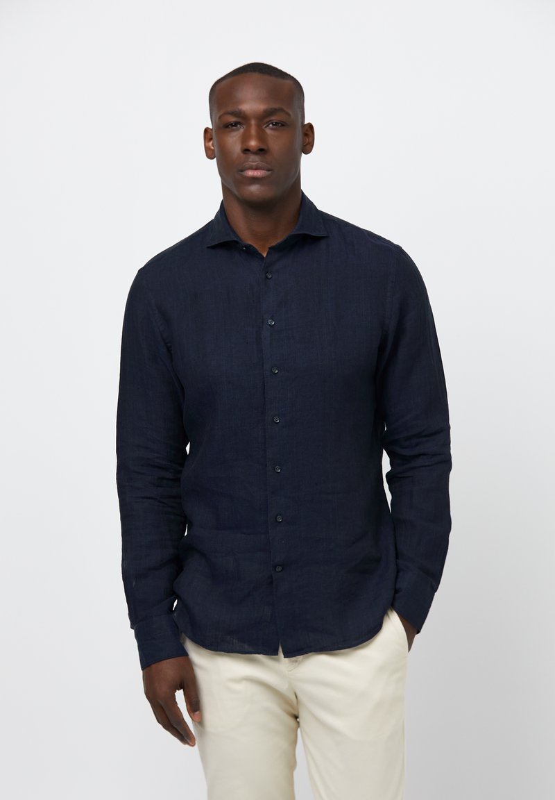 Camisa de lino azul marino con mangas largas, cierre de botones en la parte delantera y cuello extendido. Se lleva con pantalones de colores claros. Textura suave, diseño clásico.