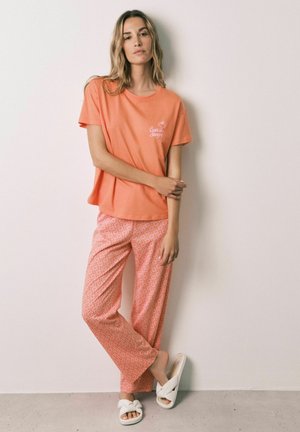 Donna con una maglietta arancione larga, pantaloni corallo fantasia e pantofole bianche, in piedi contro una parete chiara e uniforme.