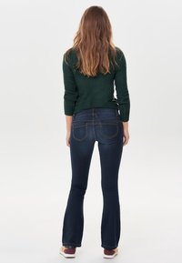 Jean flare bleu foncé à texture lisse, avec deux poches arrière et des coutures contrastées, porté avec un pull vert foncé.