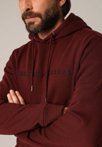 Bordeaux hoodie met een zachte textuur, voorzien van een trekkoordkap en geborduurde tekst "DELUXE ORIGINAL" in het zwart, ribbelboorden en -zoom.