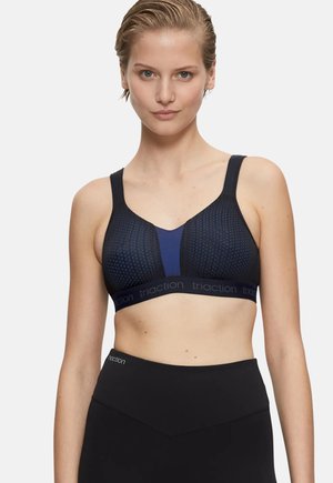 Femme aux cheveux blonds courts, portant un soutien-gorge de sport bleu marine et noir et un legging noir taille haute sur un fond uni.