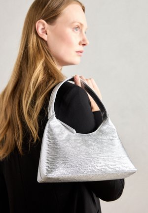 Femme aux longs cheveux blonds vêtue de noir, tenant un sac à main argenté texturé sur son épaule, sur un fond clair uni.