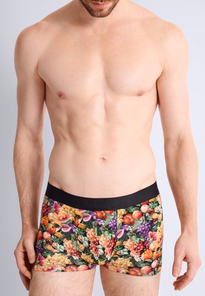Homme torse nu portant un boxer avec un motif coloré de fruits, debout devant un fond clair uni, les mains détendues le long du corps.