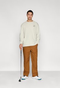 Light beige long-sleeve shirt with logo on chest, brown pants, and white golf shoes with blue accents. Standing pose, holding a golf club.