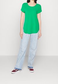 Grünes T-Shirt mit kurzen Ärmeln und rundem Ausschnitt, kombiniert mit hellblauen, locker sitzenden Jeans und burgunderroten Turnschuhen.