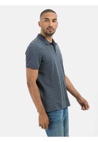 Granatowa koszulka polo wykonana z fakturowanej tkaniny, z kołnierzykiem, krótkimi rękawami i prostym krojem. Model pozuje w luźnej stylizacji w jasnych dżinsach.