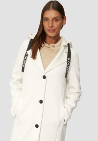 Cappotto bianco con cappuccio, accenti neri e nastri con logo. Presenta chiusura con bottoni, tasche laterali e un design strutturato con una texture morbida.