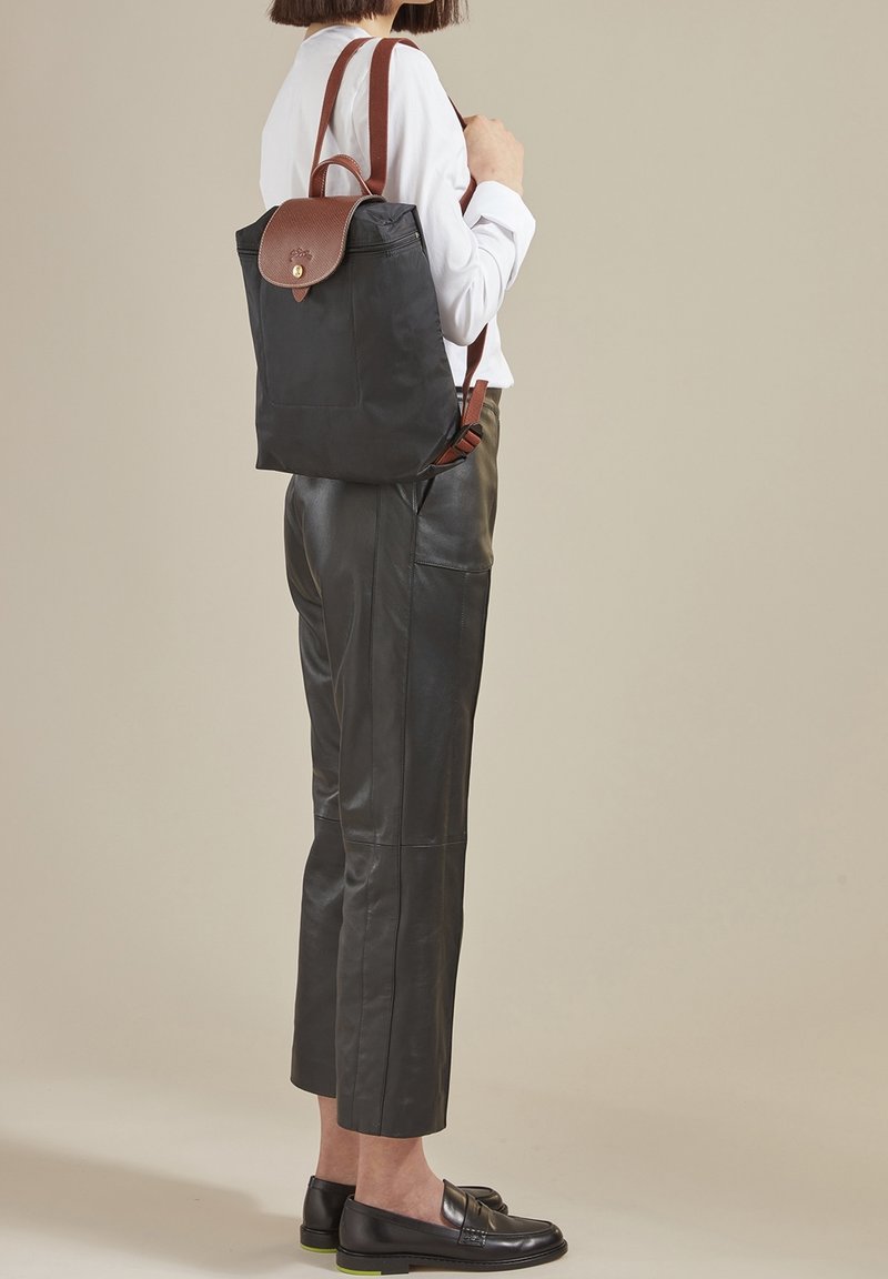 Frau trägt schwarze Leder-Cropped-Hosen, schwarze Slipper, ein weißes Langarmhemd und trägt einen schwarzen Rucksack mit braunem Lederklappen und Riemen.