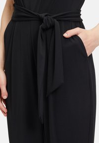 Tuta nera con cintura in tessuto annodata in vita, texture liscia e tasche laterali per una maggiore funzionalità.