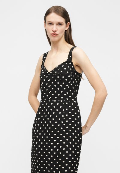 Νεαρή γυναίκα που φοράει ένα μαύρο αμάνικο φόρεμα με μοτίβα λευκών καρδιών, στέκεται με το ένα χέρι πίσω από την πλάτη της.