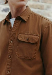 Hnedá košeľa na gombíky vyrobená z mäkkej tkaniny, s klasickým golierom, vreckom na hrudi a predným gombíkovým zapínaním s kontrastnými gombíkmi.