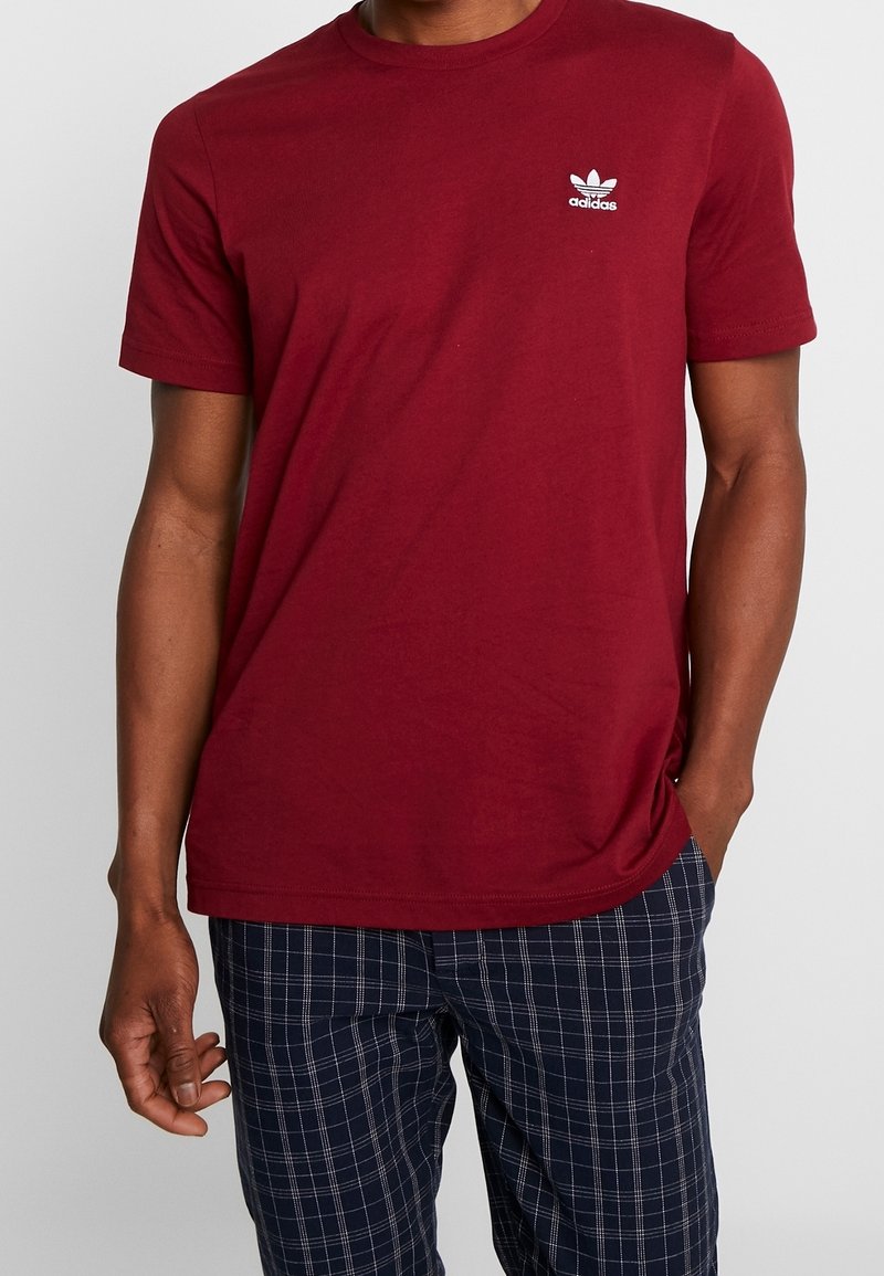 Camiseta de algodón color marrón con cuello redondo y mangas cortas. Presenta un logo de Adidas bordado en blanco en el lado izquierdo del pecho. Diseño simple.