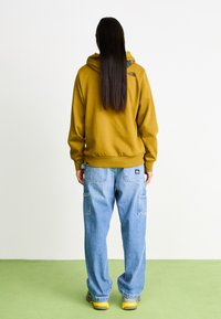 Yellow hoodie with a front pocket and logo at the back, paired with loose blue cargo jeans featuring back pockets and tags. Grey sneakers.