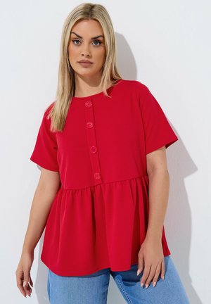 Kvinde med lyst, glat hår iført en rød kortærmet knapbluse og lyseblå jeans mod en enkel hvid baggrund.