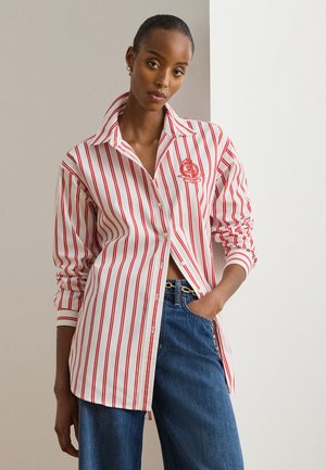 Naine, kes kannab valget punaste vertikaalsete triipudega nööbitavat särki tikitud vapiga, siniseid teksapükse ja kuldset ketivööd.