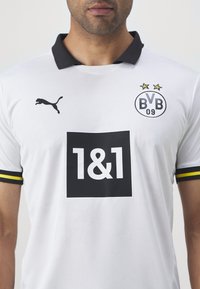 Bela nogometna majica BVB z črnim ovratnikom, ki vsebuje črn pravokotni logotip s "1&1" in črn logotip Puma na levem prsnem delu.