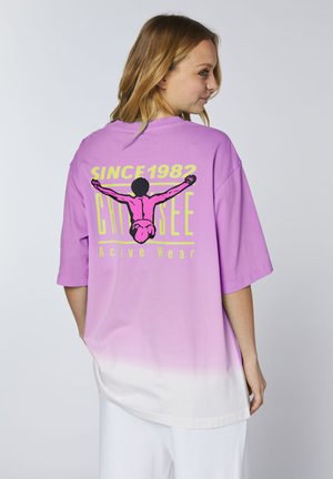 Frau trägt ein lila-weißes verlaufendes Oversize-T-Shirt mit einer Grafik eines Tauchers und dem Text „SINCE 1982 CHIYSEE Active Wear“ auf dem Rücken.