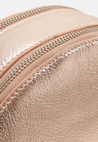 Plecak z imitacji skóry w kolorze różowego złota z dwoma metalowymi suwakami, teksturowaną powierzchnią i zaokrąglonym kształtem. Dodatki obejmują gładki wykończenie suwaka.