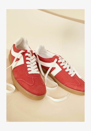 Rode suède sneakers met witte accenten, met een klassieke vetersluiting en een getextureerde gumzool. De veters zijn losgeknoopt en plat neergelegd.