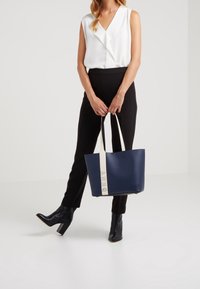 Marinblå tygväska med en strukturerad design, krämfärgade handtag med präglingen "DKNY", slätt material och en rymlig interiör.