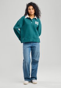 Felpa verde acqua con colletto grigio caratterizzata da un numero bianco "89," abbinata a jeans azzurri e scarpe da ginnastica bianche. Capelli ricci in vista.