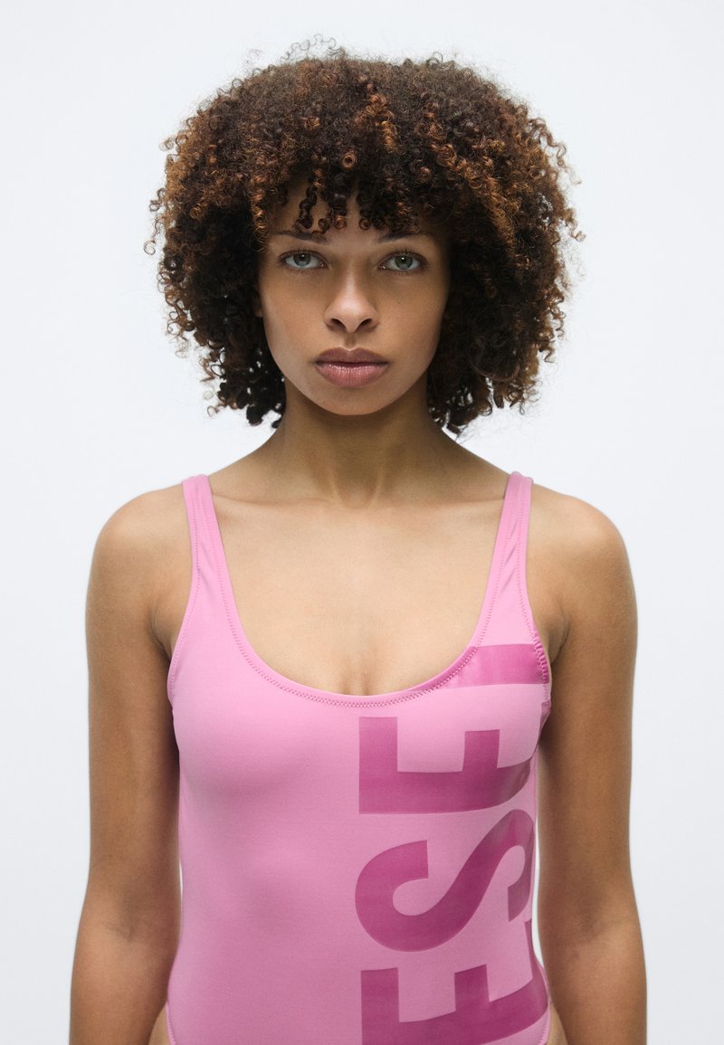 Roze badpak met een aansluitend ontwerp, voorzien van een groot gedrukt logo in een donkerdere tint roze en dunne schouderbandjes.