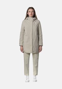 Cappotto impermeabile beige chiaro con colletto alto e zip, abbinato a pantaloni chiari semplici e sneaker bianche, indossato da una persona in piedi dritta.