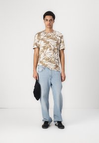 Lysebrunt t-shirt med hvidt bladmønster, parret med løse lyseblå jeans og sorte sneakers, mens der holdes en sort taske.