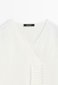 Vêtement blanc à col en V avec un volant vertical et huit petits boutons sur le devant, portant une étiquette de marque noire "Massimo Dutti".