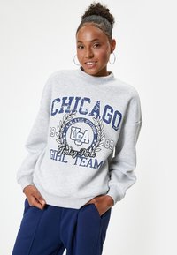 Sudadera gris con texto gráfico azul marino "CHICAGO" y "GIRL TEAM", con un escote redondo y puños de canalé. Combinada con pantalones azul marino.