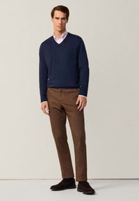 Maglione a V blu navy, camicia rosa chiaro, pantaloni marroni strutturati e scarpe scure. Sfondo semplice, posa in piedi.