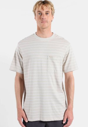 Homme blond portant un T-shirt à manches courtes rayé beige clair avec une poche poitrine, debout devant un fond blanc uni.