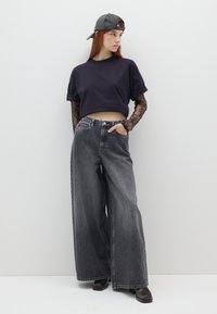 Schwarzes, kurzes T-Shirt, weit geschnittene graue Jeans, durchsichtige Spitzenärmel, schwarze Lederkappe, klobige schwarze Ankle Boots, minimaler Hintergrund.