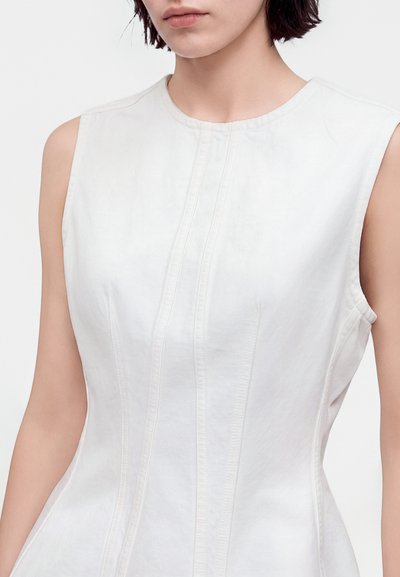 Femme portant une robe blanche sans manches avec un détail de couture verticale, cadrée sous le menton, présentée sur un fond uni.
