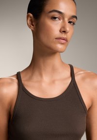 Rjava rebrasta majica brez rokavov s tankimi naramnicami, ki se tesno prilega telesu in ima gladko teksturo. Poudarite detajle izreza in ramen.