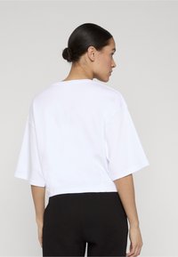 Mujer con el cabello oscuro recogido en un moño, que lleva una camiseta blanca lisa de manga corta y grande, y pantalones negros acanalados, de perfil frente a un fondo liso.