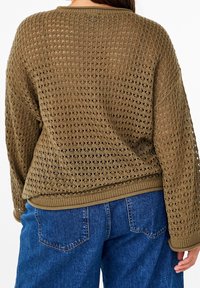 Oliwkowy sweter dzianinowy z ażurowym wzorem, luźny krój i ściągacz na dole. Połączony z niebieskimi dżinsami, z widokiem z tyłu.