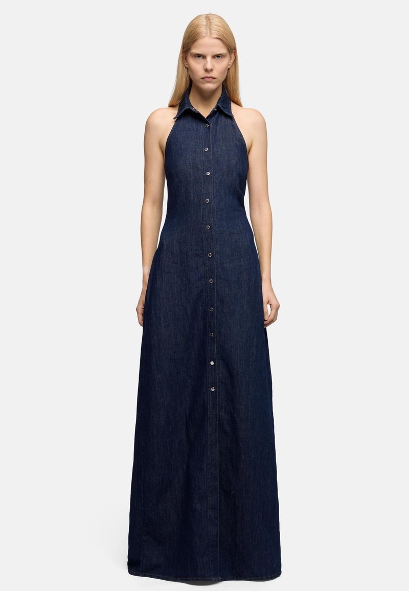 Kobieta ubrana w bezrękawnikową, ciemnoniebieską sukienkę maxi z denimu z dekoltem halter i guzikami z przodu, stojąca na tle jasnego, jednolitego tła.