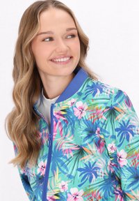 Kolorowa baza bomberka w kwiatowy wzór, z niebieskim kołnierzem, zapięciem na zamek oraz tropikalnym motywem liści w odcieniach zieleni, różu i niebieskiego.