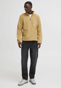 Fleece pullover in mustard yellow with a zip collar and a front pocket, paired with dark jeans and grey sneakers.