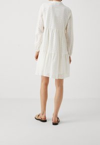 Weißes Spitzenkleid mit langen Ärmeln, gestuftem Design und rundem Ausschnitt. Der Stoff hat ein strukturiertes Muster mit Ausschnitten.