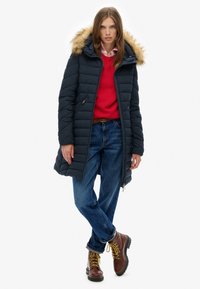 Marineblå pufferjakke med en faux fur-hette, lynlåslukninger og quiltet tekstur, båret over en rød sweater og blå jeans med snørebåndsbrune støvler.
