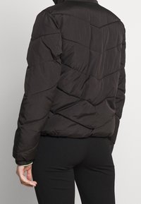Brun vadderad jacka med zigzag-quiltat mönster och hög krage. Tillverkad av ett slätt, strukturerat tyg. Har elastiska ärmslut och en figurnära design.