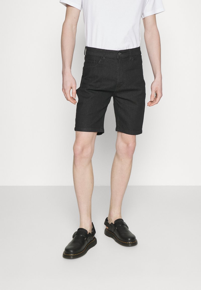 Shorts de mezclilla negra con corte ajustado, diseño de cinco bolsillos, cinturilla clásica y ligeras decoloraciones. Combinados con zapatos negros de slip-on y una camiseta blanca.