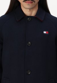 Uomo che indossa un cappotto blu navy con bottoni e colletto, con il logo di Tommy Jeans sul petto, visibile dalle spalle fino alla parte superiore del torso.