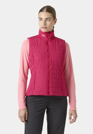 Kobieta o jasnej karnacji i związanych włosach, ubrana w jasno różową pikowaną kamizelkę na różowej, długorękawowej koszuli oraz ciemnych spodniach, patrząca w lewo.